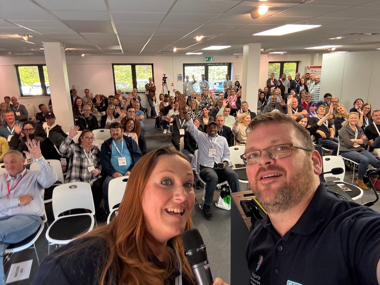 Phil and Diana on stage with a microphone in front of a packed room of instructors